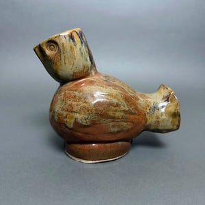 May include: A ceramic bird figurine with a glossy, multi-colored glaze featuring swirling patterns of brown, tan, and hints of blue. The sculpture has a rounded body, a short tail, and a stylized head with an etched eye, resting on a circular base.