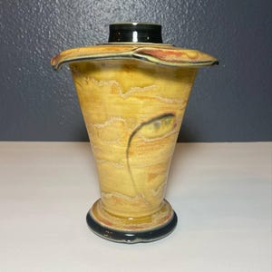 May include: A handcrafted ceramic vase with a flared rim and a black top. The vase features a yellow and brown color scheme with a unique, organic design. The base is black with a red and yellow accent.