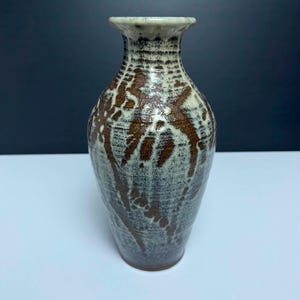 May include: A ceramic vase with a narrow neck and a bulbous body, featuring a mottled glaze in shades of cream, gray, and dark brown. The surface displays a textured, layered appearance with dark brown abstract markings over a lighter base.
