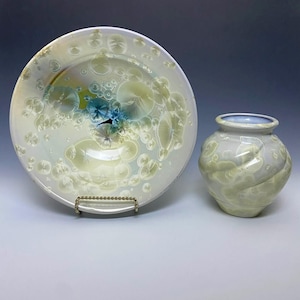 May include: A ceramic bowl and vase set. The bowl is white with a light green and blue crystalline pattern. The vase is a similar color and pattern, with a rounded shape and a light blue rim. The bowl is on a metal stand.