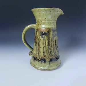 May include: Handmade ceramic pitcher with a wide spout and a curved handle. The pitcher has a light green base color with brown and black abstract patterns. The pitcher is approximately 8 inches tall.