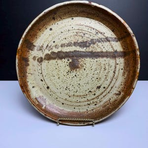 May include: A round ceramic plate with a speckled cream-colored center and a wide, textured brown rim. The surface features dark brown horizontal brushstrokes and scattered dark speckles throughout, with a slightly uneven, hand-crafted edge.