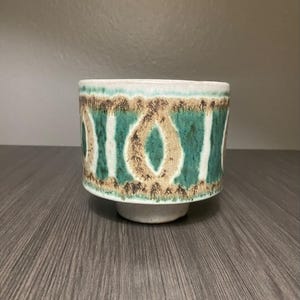 May include: A white ceramic planter with a green and brown geometric pattern. The planter has a wide base and a slightly flared rim.