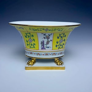 May include: A decorative porcelain bowl with a wide, flared rim and a yellow exterior. The bowl features floral and vine designs in green, purple, and white. It rests on a square base with gold-colored claw feet and trim.