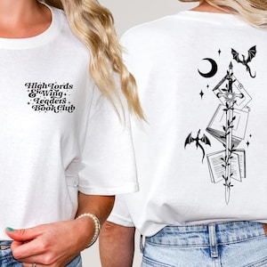 Könnte beinhalten: Weißes T-Shirt mit schwarzen Grafikdesigns. Auf der Vorderseite steht der Text "High Lords & Wingless Book Club". Auf der Rückseite befindet sich ein Design aus Büchern, Drachen, einem Schwert, einem Halbmond und Sternen. Das Shirt ist ein lässiger Unisex-Stil.