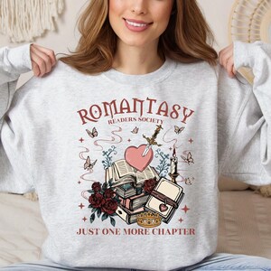 Peut inclure: Sweat-shirt gris clair avec l'inscription "ROMANTASY READERS SOCIETY" et "JUST ONE MORE CHAPTER". Le motif comprend un cœur transpercé par une épée, des livres, des papillons, des roses et une couronne. Un sweat-shirt de style décontracté et confortable.