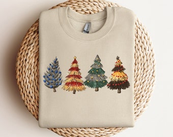 Puede incluir: Sudadera beige con cuatro árboles de Navidad bordados. Los árboles son azules, rojos, verdes y marrones, decorados con adornos y una estrella. La sudadera está doblada sobre una superficie circular tejida.