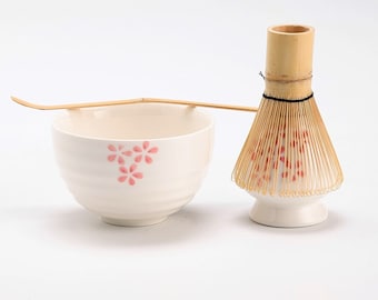 May include: A white ceramic matcha bowl with a pink floral design, a bamboo matcha whisk with a white ceramic base, and a bamboo matcha scoop.