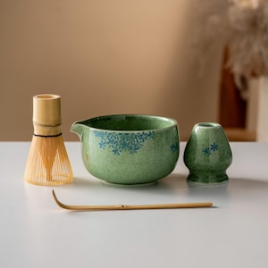 May include: A matcha tea set comprising a green ceramic bowl with blue floral patterns, a bamboo whisk, a small green ceramic tea caddy, and a bamboo scoop. Ideal for preparing and enjoying matcha tea.