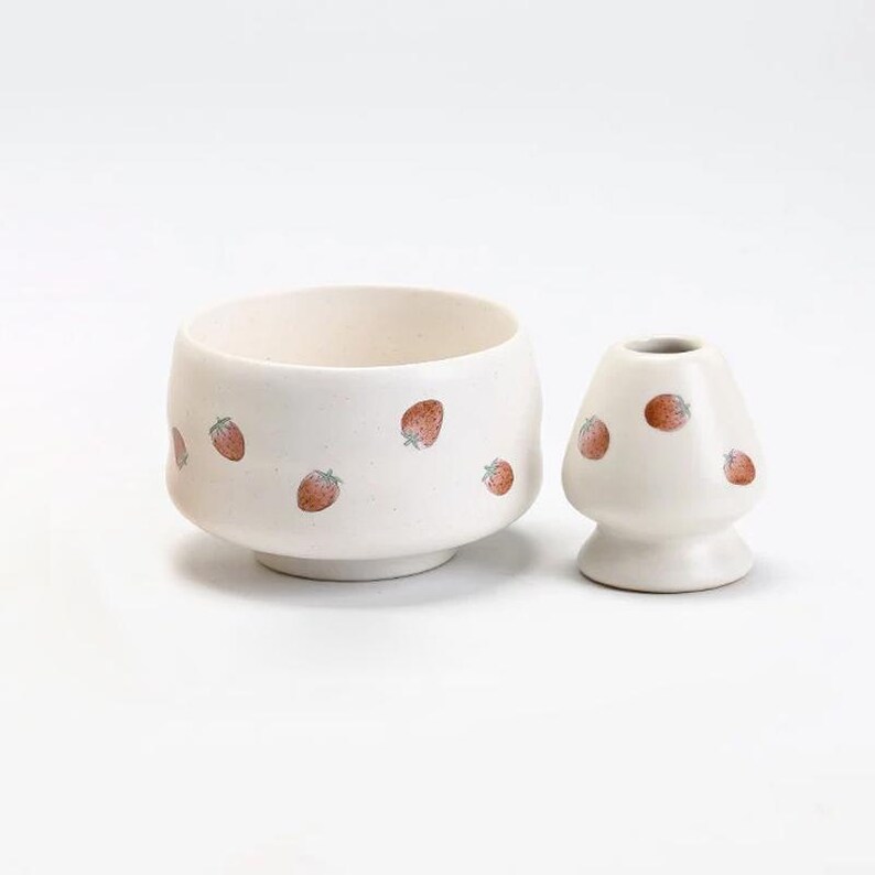 K&ouml;nnte beinhalten: Wei&szlig;es Keramik-Matcha-Set mit einem Erdbeermuster. Die Sch&uuml;ssel hat einen breiten, abgerundeten Boden und einen leicht ausgestellten Rand. Der Schneebesen ist ein traditioneller Bambusschneebesen mit einem langen Griff.