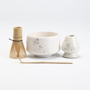 以下が含まれることがあります： 抹茶ティーセット。セラミック製のボウル、泡立て器、泡立て器ホルダー、竹製のスクープが含まれています。オフホワイトのセラミック作品には、漫画の犬とピンクの花柄のアクセントが施されています。泡立て器は竹製です。