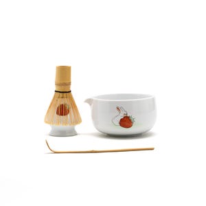May include: A white ceramic matcha bowl with a hand-painted design of a white rabbit eating a strawberry. The bowl is accompanied by a bamboo matcha whisk and a bamboo matcha scoop.
