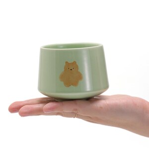 May include: A light green ceramic teacup with a brown bear design. The cup is sitting on a hand.