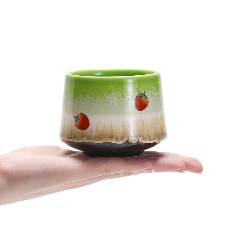 May include: A green ceramic teacup with a brown rim and two red strawberry designs. The cup is being held in a hand.