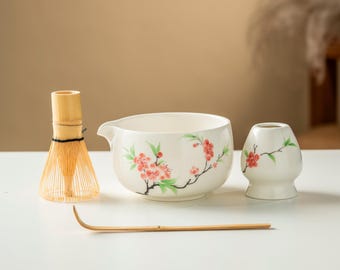 May include: A matcha tea set featuring a bamboo whisk, a ceramic bowl with a spout, a small ceramic holder, and a bamboo scoop. The white ceramic pieces are decorated with red and green floral designs. The whisk is made of light-colored bamboo.