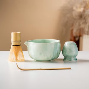 Puede incluir: Un juego de té matcha con un cuenco de cerámica verde claro, un batidor, un jarrón pequeño y una cuchara de bambú. El cuenco tiene un pico. El juego está dispuesto sobre una superficie blanca.