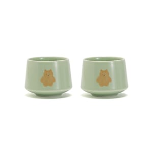 May include: Two light green ceramic teacups with a brown bear design. The cups have a rounded bottom and a slightly flared lip.