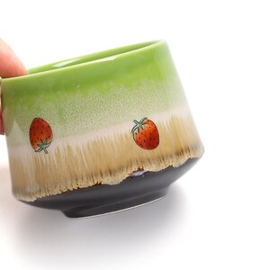 May include: A green ceramic teacup with a brown rim and two red strawberry designs. The cup has a speckled glaze and a light brown band.