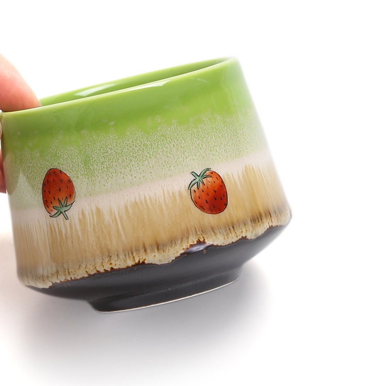 May include: A green ceramic teacup with a brown rim and two red strawberry designs. The cup has a speckled glaze and a light brown band.