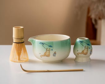 May include: A matcha tea set featuring a bamboo whisk, a ceramic bowl with a green ombre design, a small ceramic holder, and a bamboo scoop. The bowl and holder are decorated with a cat illustration. The set is designed for preparing matcha tea.