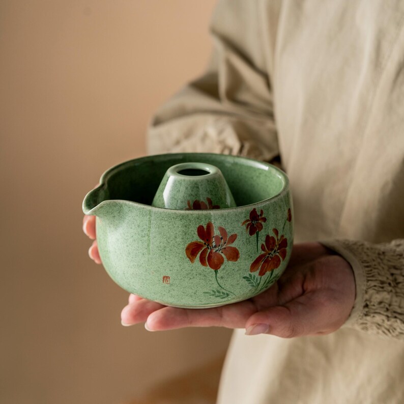May include: A green ceramic tea bowl with a central cone-shaped feature and a spout. The bowl is decorated with red floral designs. The tea bowl is held in a person's hands.