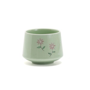 May include: A light green ceramic teacup with a floral design. The cup has two pink flowers with green stems and leaves.