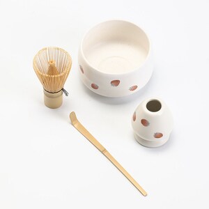 K&ouml;nnte beinhalten: Eine wei&szlig;e Keramik-Matcha-Schale mit Erdbeermuster, ein Bambus-Schneebesen, ein Bambus-Matcha-L&ouml;ffel und eine wei&szlig;e Keramik-Teetasse mit Erdbeermuster.