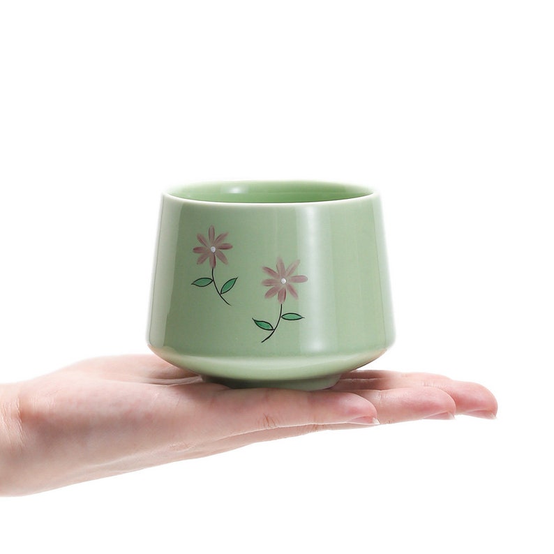 May include: A light green ceramic teacup with a floral design. The cup has two pink flowers with green stems and leaves.