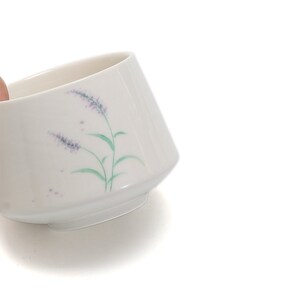 以下が含まれることがあります： 紫と緑の花が描かれた白い陶器のボウル。ボウルは縁が少し広がっていて、丸い底をしています。