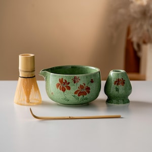 Puede incluir: Un juego de té matcha con un cuenco de cerámica verde con un pico y un diseño floral rojo, un batidor de bambú, un pequeño soporte de cerámica verde y una cuchara de bambú. El juego está sobre una superficie blanca.
