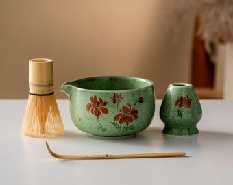 Może przedstawiać: Zestaw do herbaty matcha z zieloną ceramiczną miską z wylewką i czerwonym kwiatowym wzorem, bambusową trzepaczką, małym zielonym ceramicznym uchwytem i bambusową łyżką. Zestaw jest na białej powierzchni.