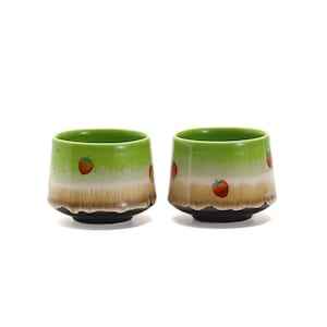 May include: Two green ceramic teacups with a brown rim and a white band with red strawberry designs.