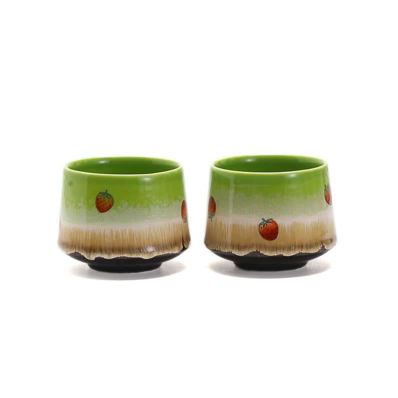 May include: Two green ceramic teacups with a brown rim and a white band with red strawberry designs.