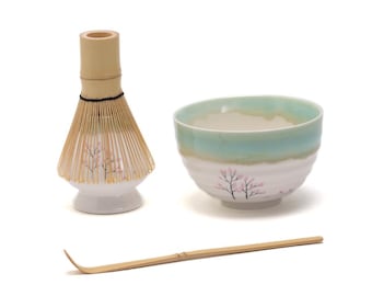 May include: A white ceramic matcha bowl with a light blue and green glaze and a floral design. A bamboo matcha whisk with a white base and a bamboo matcha scoop are also included.