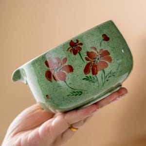 May include: A green ceramic bowl with a speckled texture and a floral design in shades of red and pink. The bowl has a curved shape with a small handle on one side. The flowers are painted with delicate details, and the bowl is held in a hand.