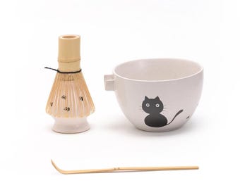 May include: A white ceramic matcha bowl with a black cat design, a bamboo whisk, and a bamboo matcha scoop.