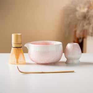 Op de afbeelding: Een matcha-theeset met een bamboe klopper, een roze en witte keramische kom, een kleine roze en witte keramische theekop en een bamboelepel. De kom heeft een tuit. De set is op een wit oppervlak geplaatst.