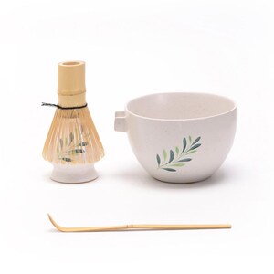 May include: A white ceramic matcha bowl with a green leaf design, a bamboo matcha whisk with a white ceramic base, and a bamboo matcha scoop.
