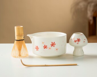 May include: A white ceramic matcha bowl, mushroom-shaped vase, bamboo whisk, and scoop. The bowl and vase are decorated with pink floral designs. The whisk is made of thin bamboo strips.