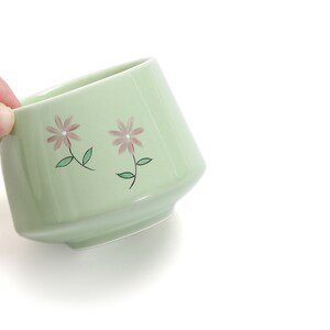 May include: A light green ceramic teacup with a floral design. The cup has two pink flowers with green leaves.