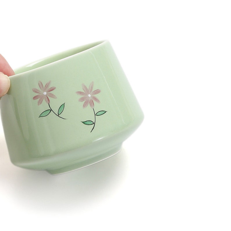 May include: A light green ceramic teacup with a floral design. The cup has two pink flowers with green leaves.
