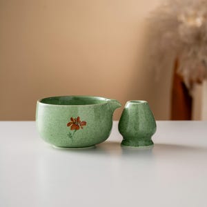 May include: A green ceramic matcha bowl with a spout and a matching whisk holder. The bowl features a hand-painted orange flower design. The items are set on a white surface, with a neutral background.