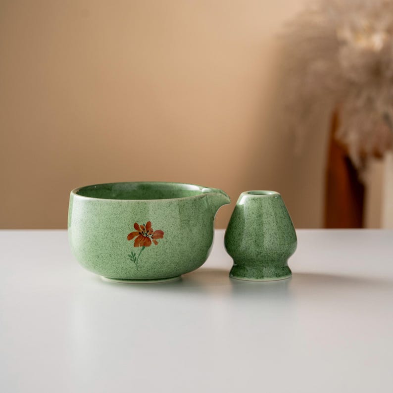 May include: A green ceramic matcha bowl with a spout and a matching whisk holder. The bowl features a hand-painted orange flower design. The items are set on a white surface, with a neutral background.