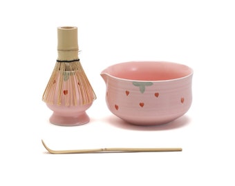 May include: A pink ceramic matcha bowl with a strawberry design, a bamboo matcha whisk with a pink base, and a bamboo matcha scoop.