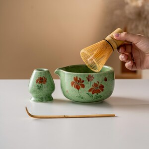 May include: A matcha tea set featuring a green ceramic bowl and a small vase, both adorned with red floral designs. A bamboo whisk and scoop are also present. The whisk is being held above the bowl. The set is designed for preparing matcha tea.