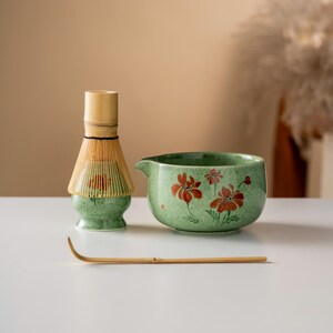 May include: A matcha tea set featuring a green ceramic bowl with red floral designs, a bamboo whisk, and a bamboo scoop. The whisk has a light brown handle and a green base. The set is designed for preparing matcha tea.