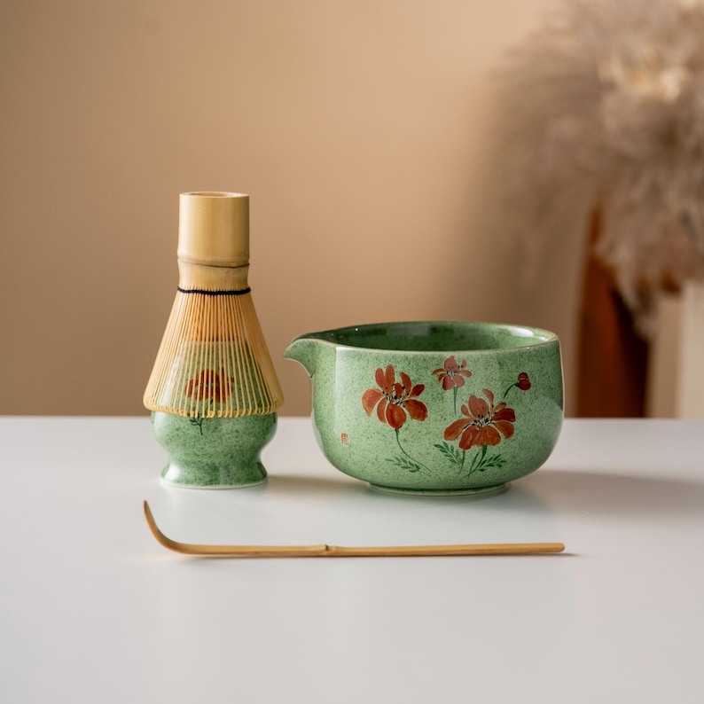 May include: A matcha tea set featuring a green ceramic bowl with red floral designs, a bamboo whisk, and a bamboo scoop. The whisk has a light brown handle and a green base. The set is designed for preparing matcha tea.