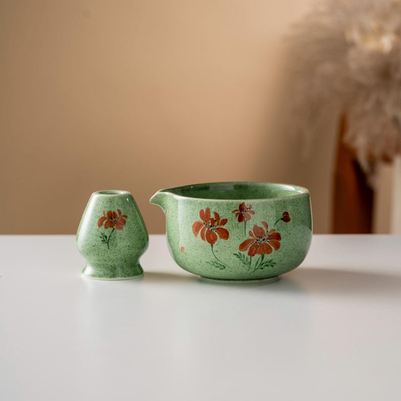 May include: A green ceramic tea bowl and whisk holder set. The set features a speckled green glaze with hand-painted red floral designs. The bowl has a spout for easy pouring. The whisk holder is a small, vase-like shape.