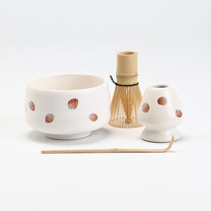 K&ouml;nnte beinhalten: Eine wei&szlig;e Keramik-Matcha-Schale mit Erdbeermuster, ein Bambus-Schneebesen, ein wei&szlig;er Keramik-Schneebesenhalter mit Erdbeermuster und ein Bambus-Matcha-L&ouml;ffel.
