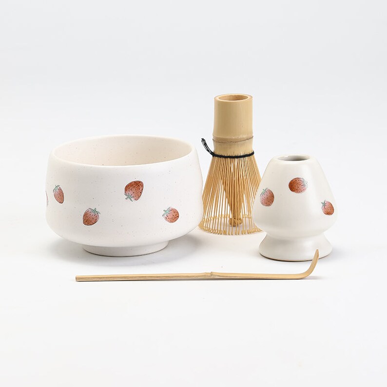 K&ouml;nnte beinhalten: Eine wei&szlig;e Keramik-Matcha-Schale mit Erdbeermuster, ein Bambus-Schneebesen, ein wei&szlig;er Keramik-Schneebesenhalter mit Erdbeermuster und ein Bambus-Matcha-L&ouml;ffel.
